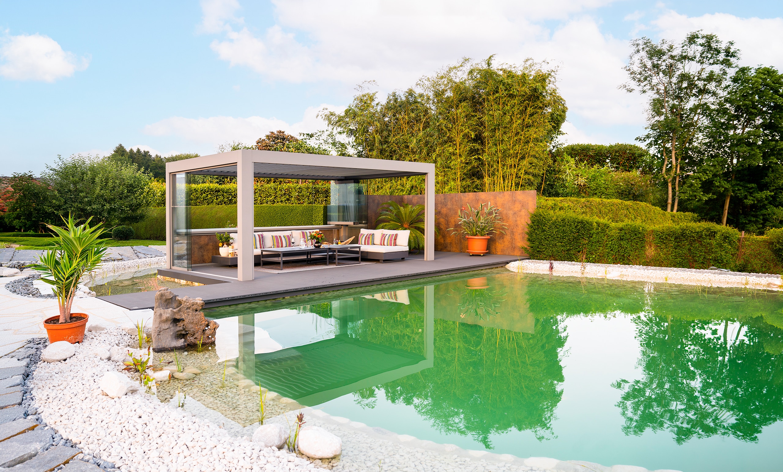 Ein moderner Garten mit Teich und hellgrauer Pergola mit halbgeöffnetem Lamellendach und offenen Glasschiebetüren von Moltke.