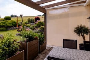 Newo Sonnen- und Insektenschutz zeigt eine überdachte Terrasse, die mit beigen Textilscreens seitlich geschlossen werden kann.