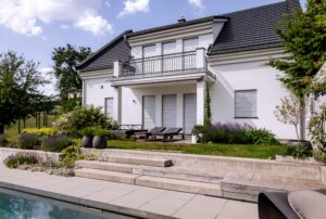 Newo Sonnen- und Insektenschutz zeigt ein Einfamilienhaus mit Terrasse und Balkon und bodentiefen Fenstern, die mit weißen Raffstores beschattet werden.