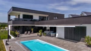 Eine Flachdachvilla mit bodentiefen Fenstern mit Außenjalousien und einem Poolbereich mit elektrischer Markise in Weiß-Grau, der Schatten spendet und das Terrassenambiente und ergänzt von Newo.