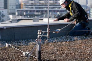 Oö. Blitzschutzgesellschaft zeigt einen Arbeiter auf einem Dach, der am Blitzschutz mit integrierter Absturzsicherung mit einem Seil gesichert ist.