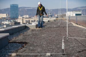 Oö. Blitzschutzgesellschaft zeigt einen Arbeiter auf einem Dach, der am Blitzschutz mit integrierter Absturzsicherung mit einem Seil gesichert ist.