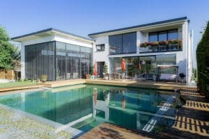Villa mit Balkon, einer Terrasse mit Holzsteg und Outdoor-Fliesen in Holzoptik, integrierten Lichtspots, Naturpool, einer Hecke mit Torbögen und großzügigem Garten von Planungsbüro Schaufler.