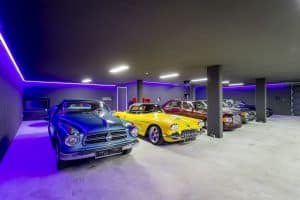 Garage im Stil eines Privatmuseums mit LED-Lichleiste zwischen Decke und Wand in Violett, rechteckigen Deckenleuchten und hellgrauem Estrich. Die Oldtimer in verschiedenen Farben sind umgeben von Säulen, die zusammen mit Decke und Wänden Anthrazit gestrichen sind von Planungsbüro Schaufler.