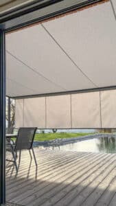 Eine Holzterrasse mit verlängerter halbtransparenter Markise in Grau bzw. Beige mit Blick auf den Naturpool von Sonnenschutz Rachbauer.