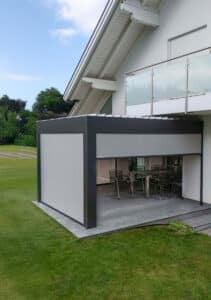 Ein Haus mit Balkon und anthrazitfarbener Pergola‑Terrassenüberdachung, halbgeöffnetem weissen Lamellendach und silbernen Senkrechtmarkisen von Sonnenschutz Rachbauer.