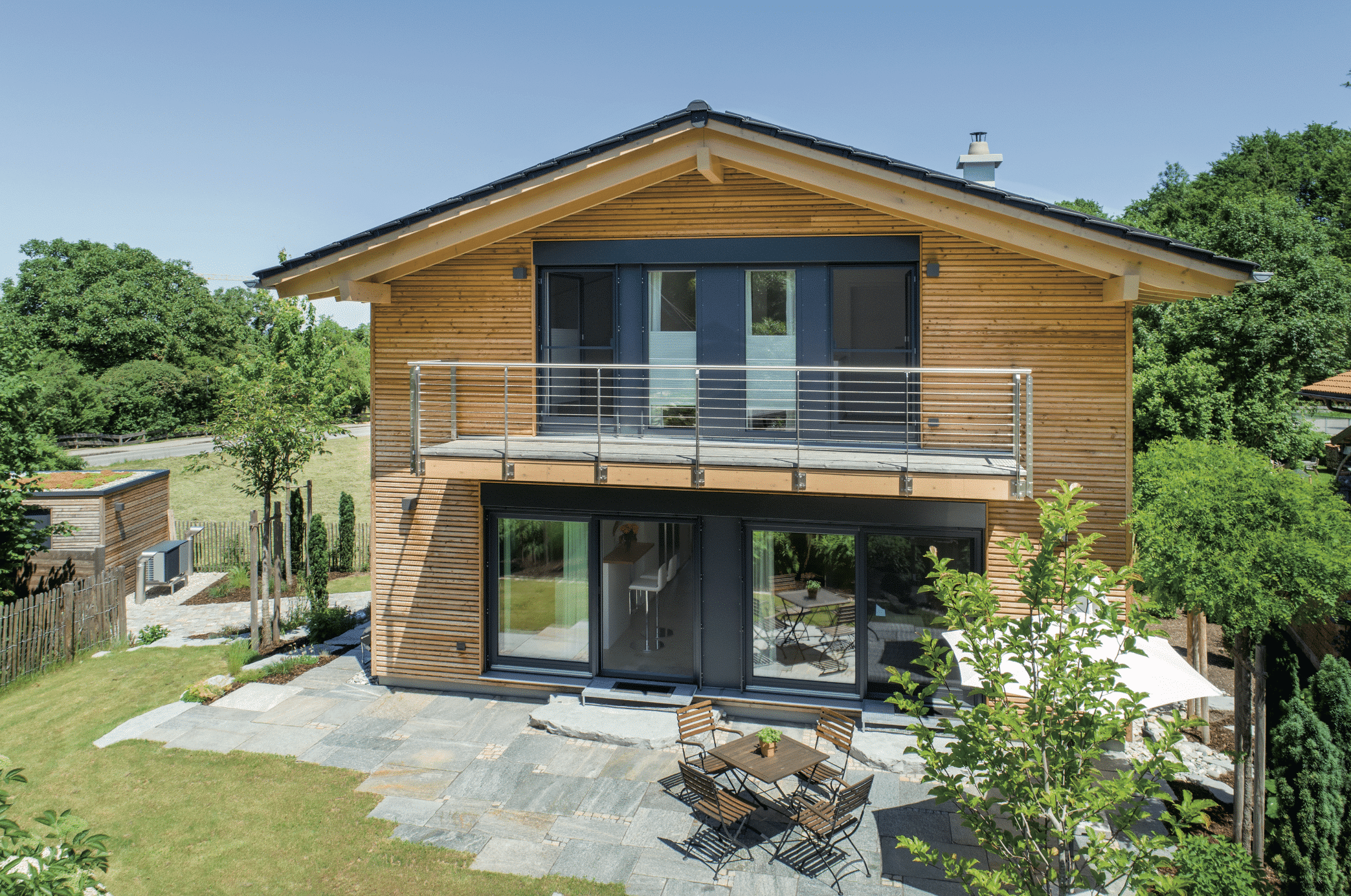 Regnauer zeigt ein rustikales Einfamilienhaus mit schöner Holzverkleidung und einer Terrasse mit grauen Steinplatten und einer dunklen Sitzgruppe auf Holz.