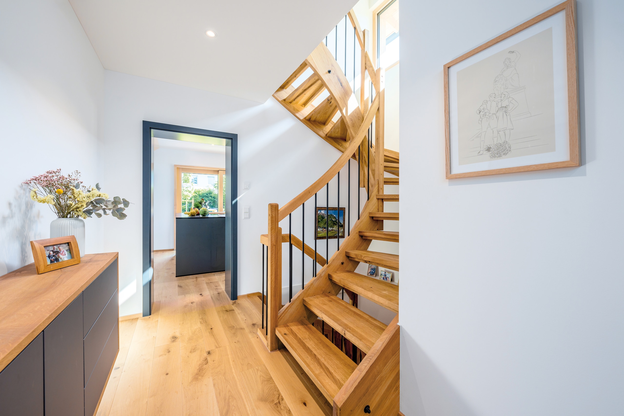Regnauer Fertighaus zeigt lichtdurchfluteten Vorraum mit geschwungener Holztreppe, Holzboden, dunkelgrauen Türrahmen und Sideboard mit Holzkorpus und dunkelgrauer Front.