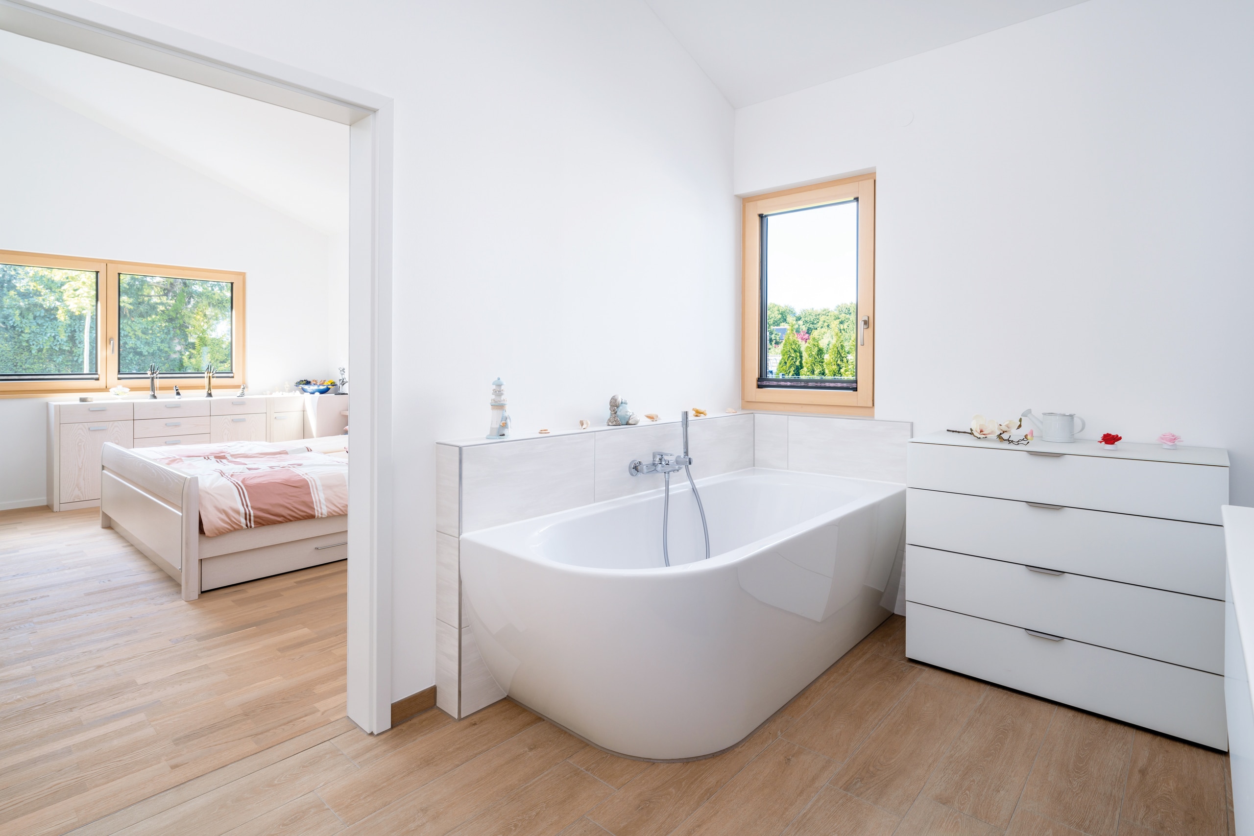 Regnauer Fertighaus zeigt helles Badezimmer mit moderner Badewanne, Holzboden, Holzrahmenfenster und Blick in das Schlafzimmer.