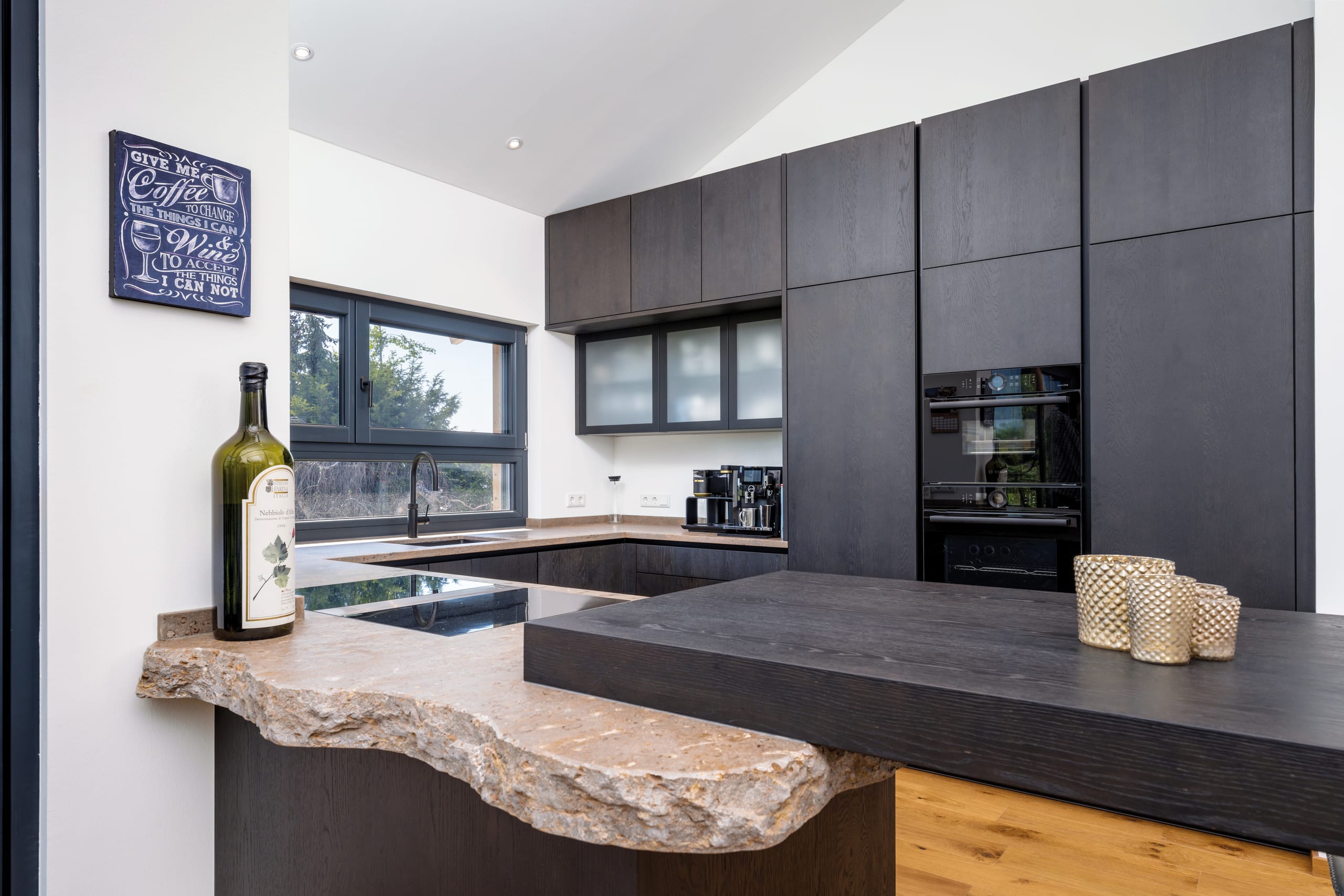 Regnauer Fertighaus zeigt elegante Küche mit dunkelgrauen Fronten, einer Steinarbeitsplatte dessen Bruchkante und angebautem Holzplatte als Bar dient.