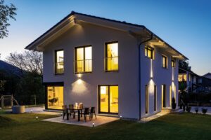Ein modernes Satteldach‑Haus bei Nacht mit auskragendem Dachvorsprung, französischem Balkon, dreiseitiger Eckverglasung, gefliester Terrasse und Gartenbrunnen von Regnauer.