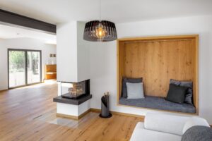 Ein offener Wohnbereich mit gemauertem Kamin, einer dreiseitigen Couch und einer gemütlichen Sitznische mit Holzverkleidung und gepolsterter Sitzfläche von Regnauer.