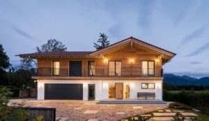 Eine Villa mit Holzfassade im Obergeschoss, überdachtem Rundumbalkon, Seitenlicht beim Hauseingang, dunkelgrauer Doppelgarage mit Sektionaltor bei Nacht – von Regnauer.