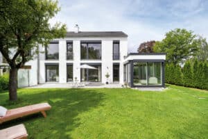Ein Fertighaus mit Flachdach angebautem Wintergarten, französischen Balkonen, Gartenbereich, hochgezogenen Raffstores und Verglasung mit schwarzem Alu‑Rahmen von Regnauer Hausbau.