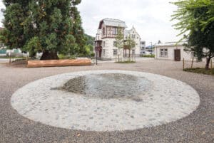 RÖFIX zeigt die Außenansicht einer renovierten Villa mit Satteldach, Fachwerkaufbau und weißem Außenputz mit Verziehrungen hinter einem großen Park mit Brunnen.