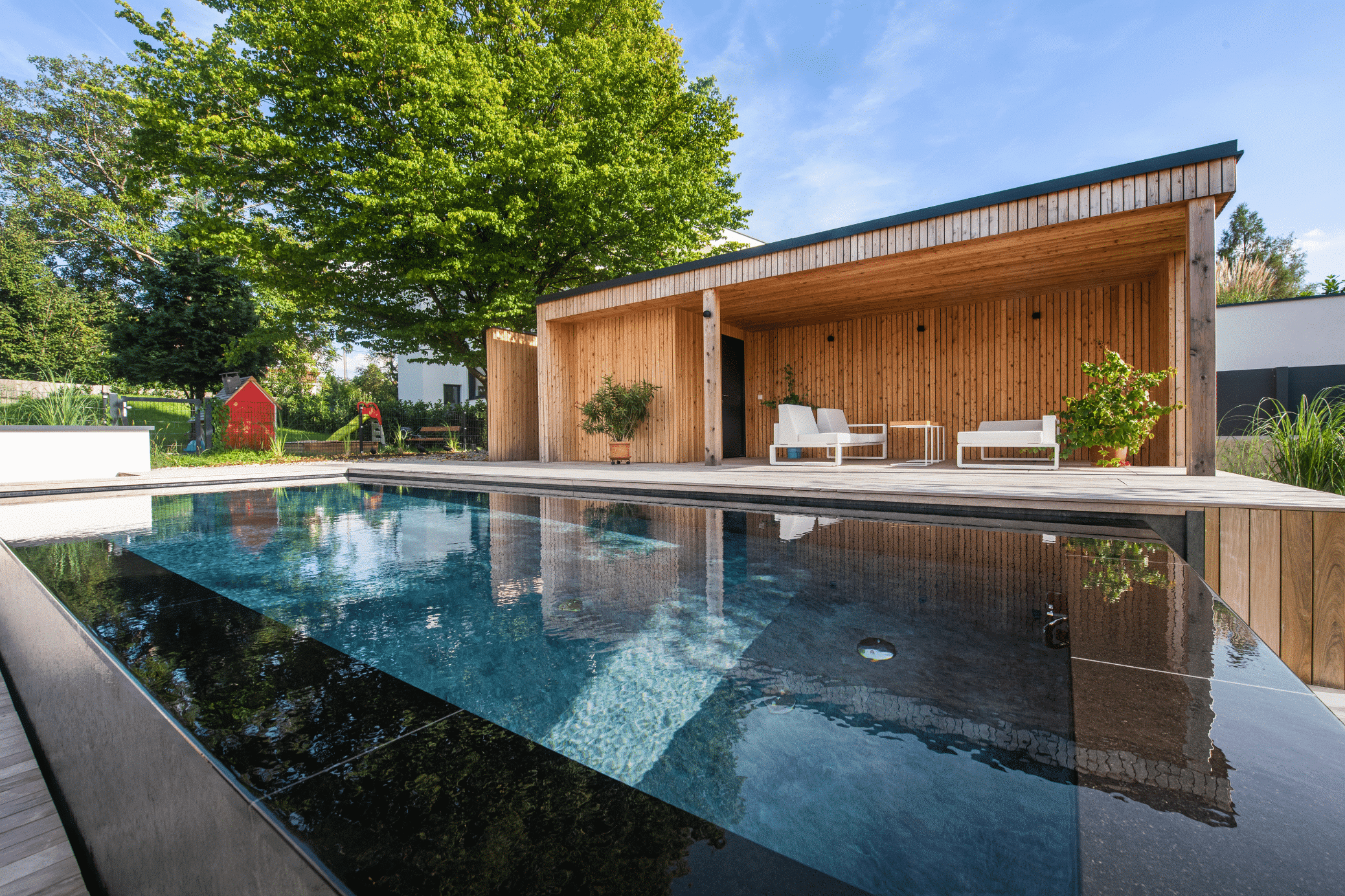 RUHA zeigt einen rechteckigen Infinity Pool mit schwarzem Rand und Holzumrandung in einem Garten mit einer Terrasse, überdachter Sitzlounge und kleinem Spielplatz für Kinder.