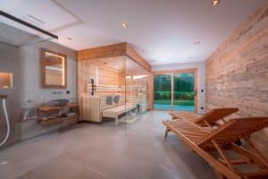 RUHA präsentiert einen Homespa-Raum mit großer Sauna aus Altholz, einem Waschbereich, einer barrierefreien Dusche und zwei Holz-Liegestühle mit Zugang auf die Terrasse.
