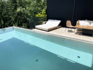 Ein Infinity‑Pool mit Überlaufrinne und Fliesenrand, Relaxliege, schwarzem Sprossen‑Terrassengeländer sowie einem Outdoortisch mit Stuhl von Ruha Stelzmüller.