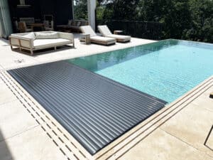 Ein Infinity‑Pool mit Überlaufrinne und Fliesen‑Poolumrandung, Relaxliege, halboffener schwarzer Poolabdeckung und schwarzem Sprossen‑Terrassengeländer von Ruha Stelzmüller.