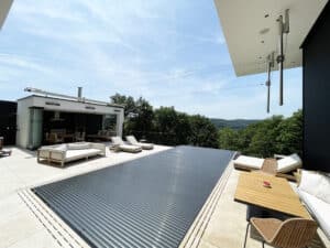 Ein Infinity‑Pool mit Überlaufrinne, Fliesenrand, Relaxliege, schwarzem Sprossen-Terrassengeländer und Poolabdeckung, daneben ein Wintergarten‑Poolhaus von Ruha Stelzmüller.