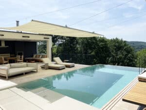 Ein Infinity‑Pool mit Kante und Überlaufrinne, Fliesenpoolumrandung, Relaxliege, Terrassengeländer in schwarzer Sprossenoptik und Sonnensegel von Ruha Stelzmüller.