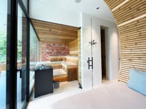 Ein Sauna­raum im Erdgeschoss mit großer Glasfront, einer bequemen Relaxliege und warmem Holzambiente von Ruha Stelzmüller.