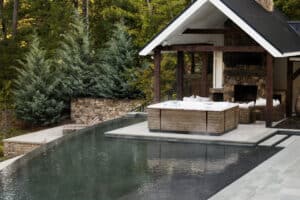 Ruha Stelzmüller zeigt ein Whirlpool, das auf einer Terrasse vor einer kleinen Hütte im Freien steht und außen mit Holz verkleidet ist.
