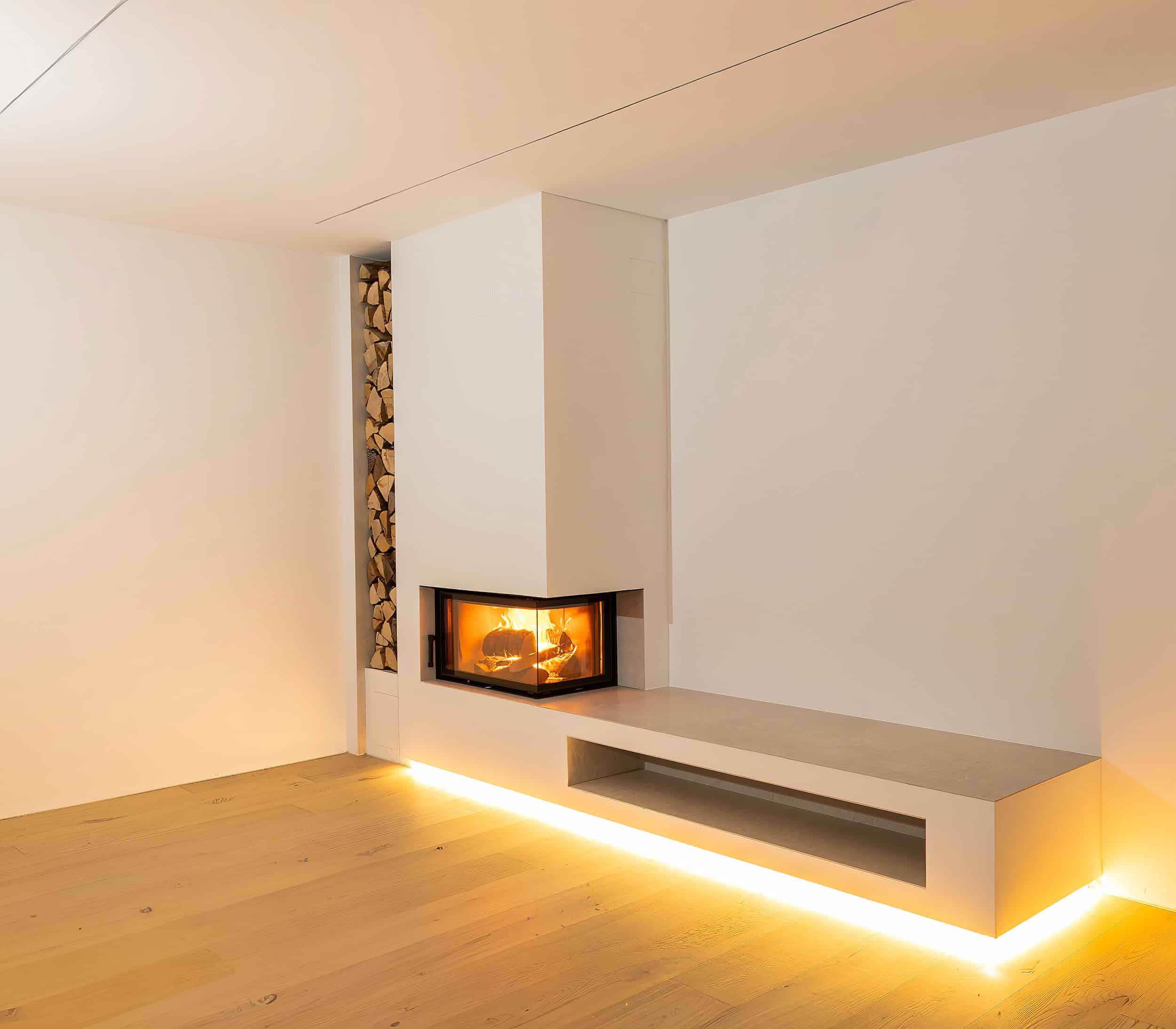 Ein moderner Kaminofen mit Holzlager und schwebender Sitzbank von unten beleuchtet präsentiert von Uwe Seidl Ofenbau.