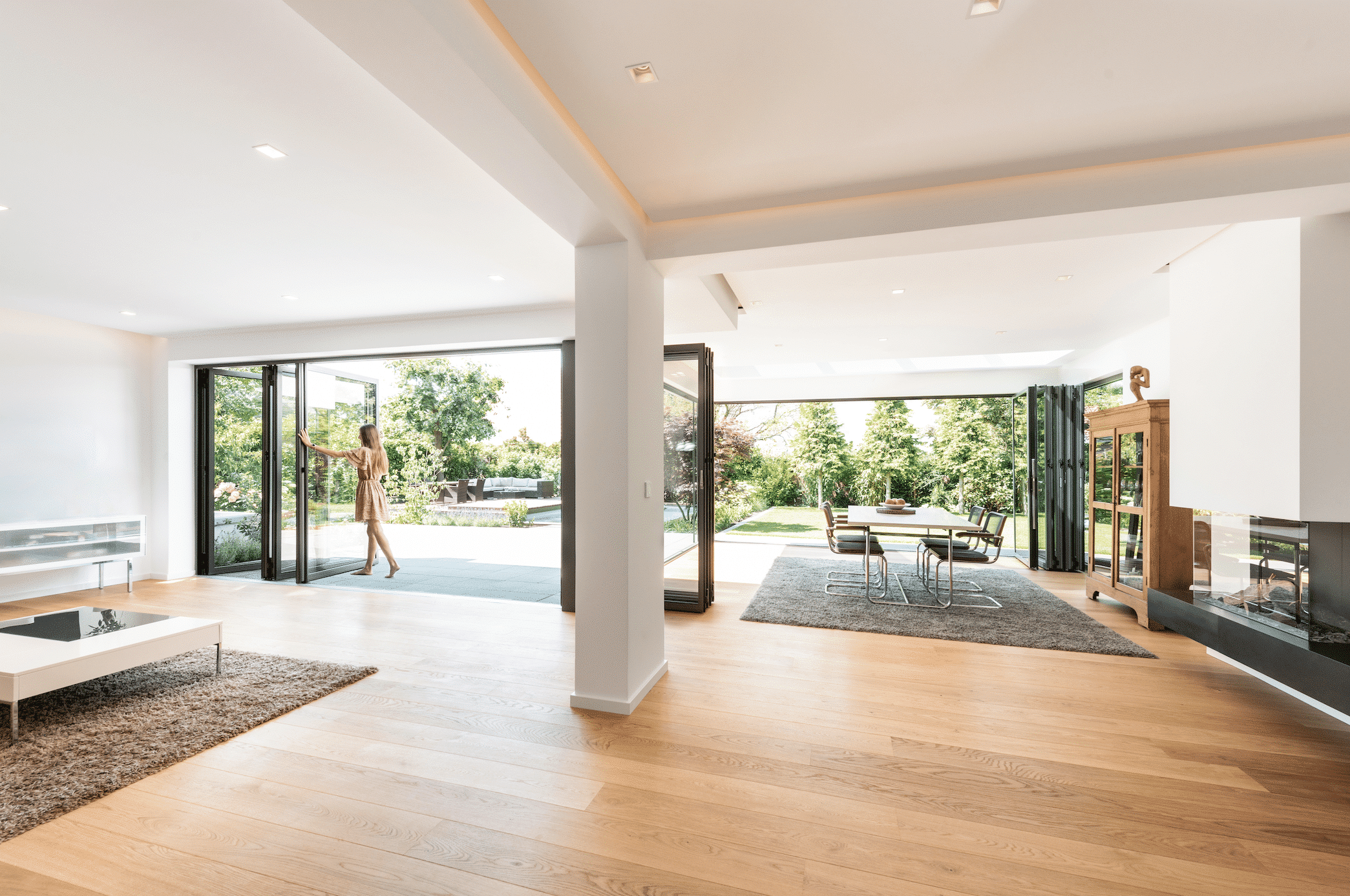 Solarlux zeigt einen hellen, offenen Wohnbereich mit einer Essgruppe, freiliegendem Teppich und der Glasfaltwand mit Zugang zum begrünten Garten mit Pool.