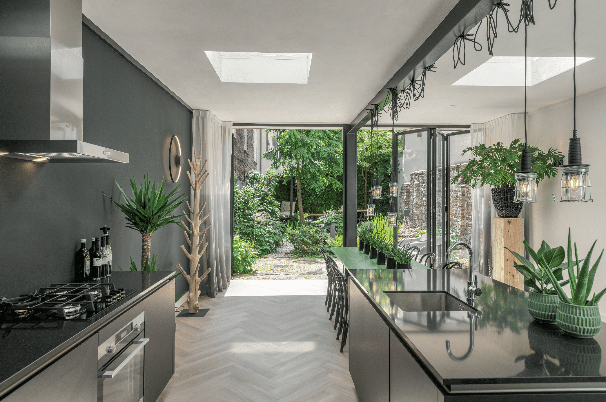 Solarlux zeigt eine moderne Küche mit dunklen Farbnuancen, vielen Pflanzen und einer Glasfaltwand mit schwarzem Rahmen und Zugang zur begrünten Terrasse.