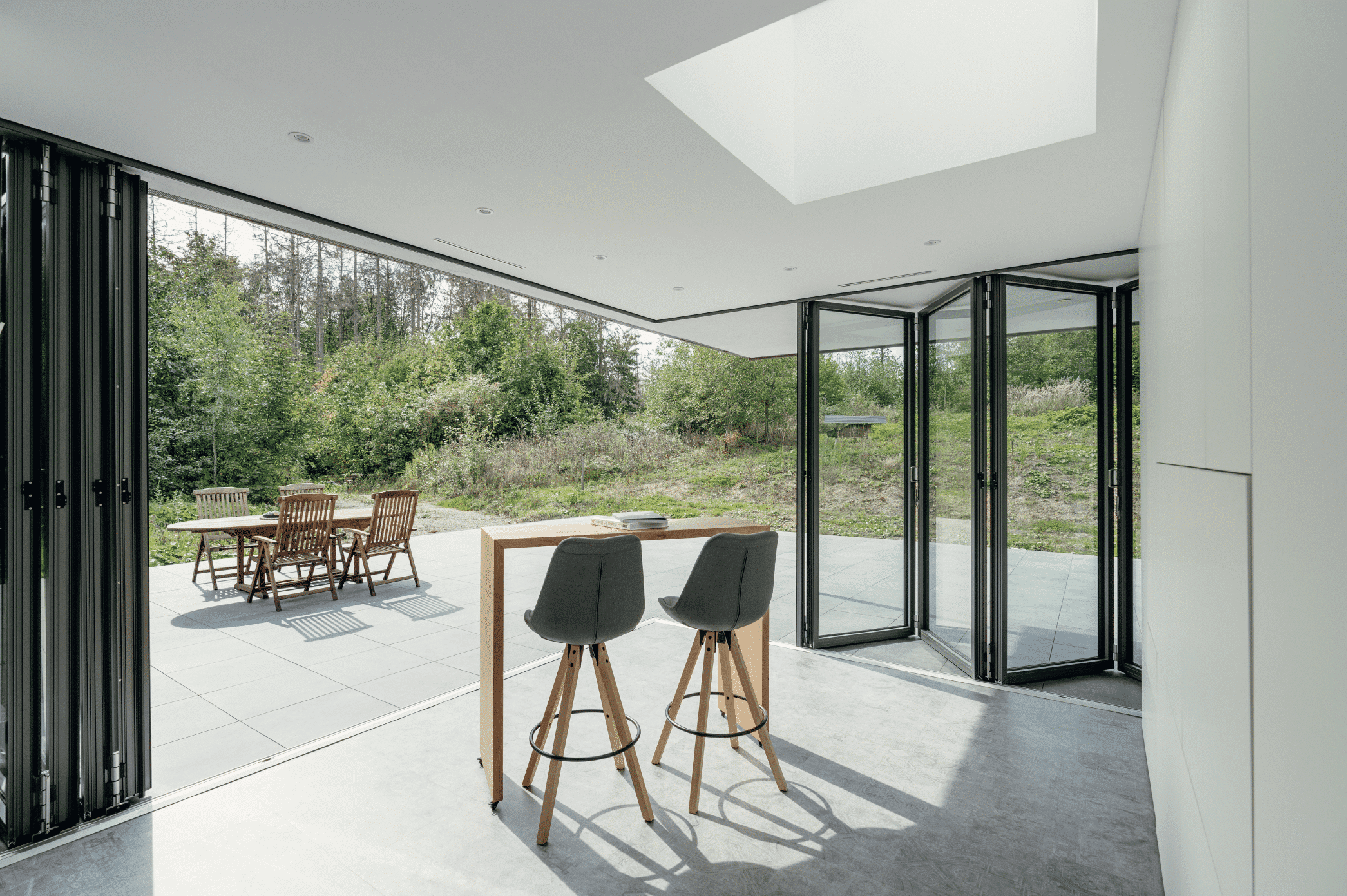 Solarlux zeigt eine geflieste Terrasse mit überdachtem Bereich mit einem Hochtisch und schwarzem Hocker der mit einer Glasfaltwand geschlossen werden kann.