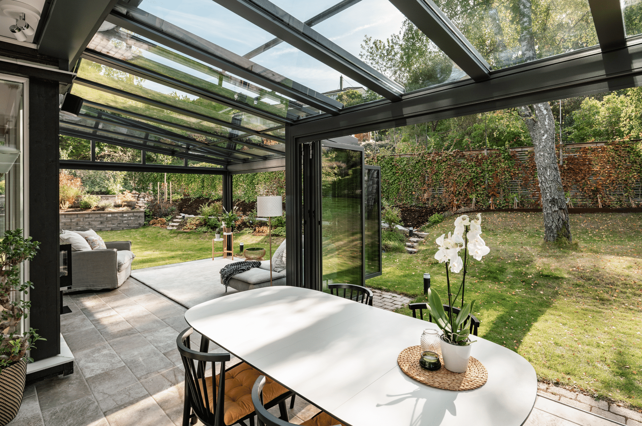 Solarlux zeigt eine große Terrasse mit Fliesenboden und Loungemöbeln und einem überdachten Bereich mit einer Sitzgruppe und Glasschiebetüren mit Zugang zum Garten.