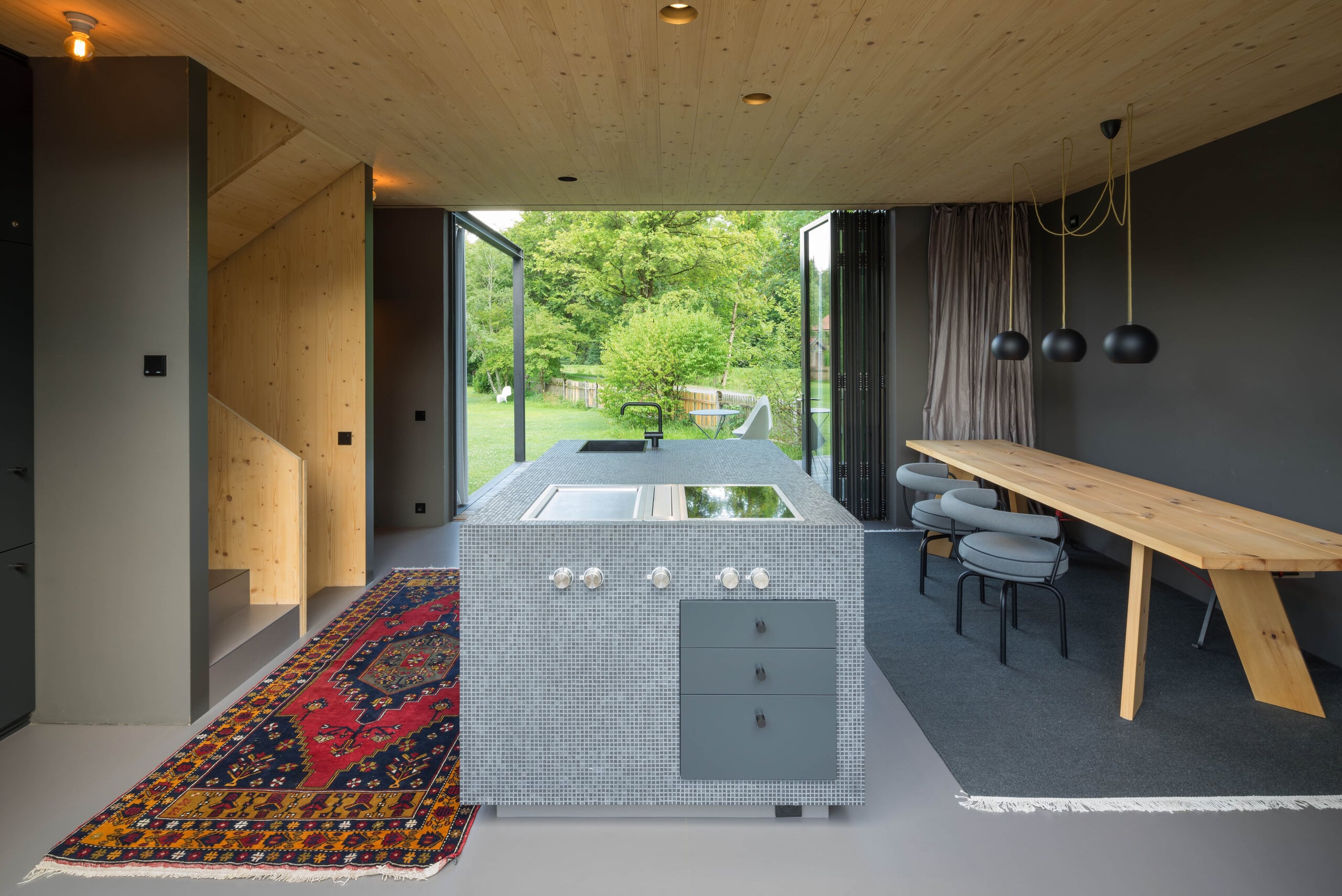 Solarlux zeigt offene Wohnküche in einem modernes Holzhaus mit geöffneten Faltschiebetüren mit dunkelgrauem Rahmen mit Blick auf den Garten.
