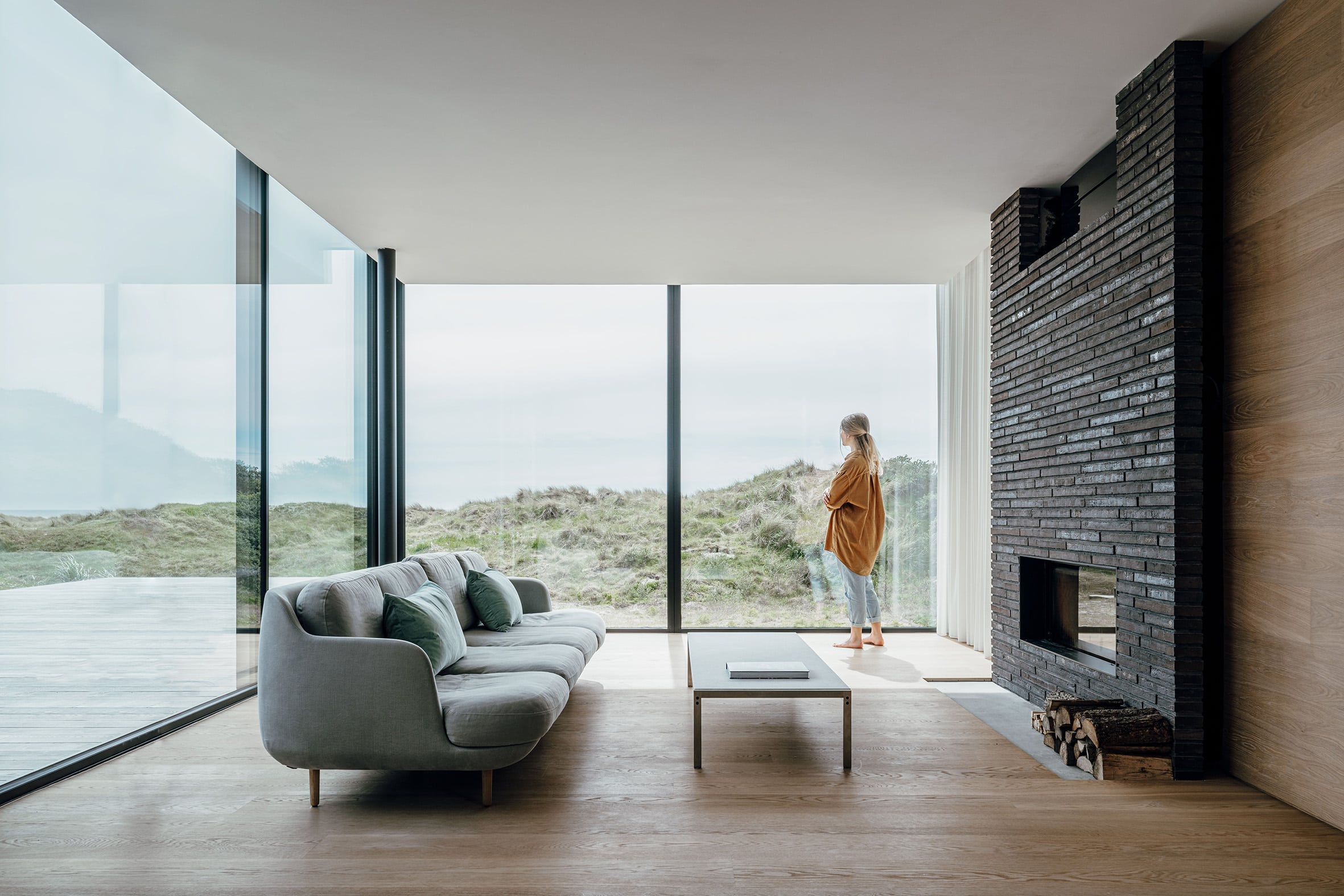 Eine Frau steht in einem großzügigem Wohnraum mit gemauertem Kamin, Sofalandschaft und Eckverglasung mit Panorama-Aussischt von Solarlux.