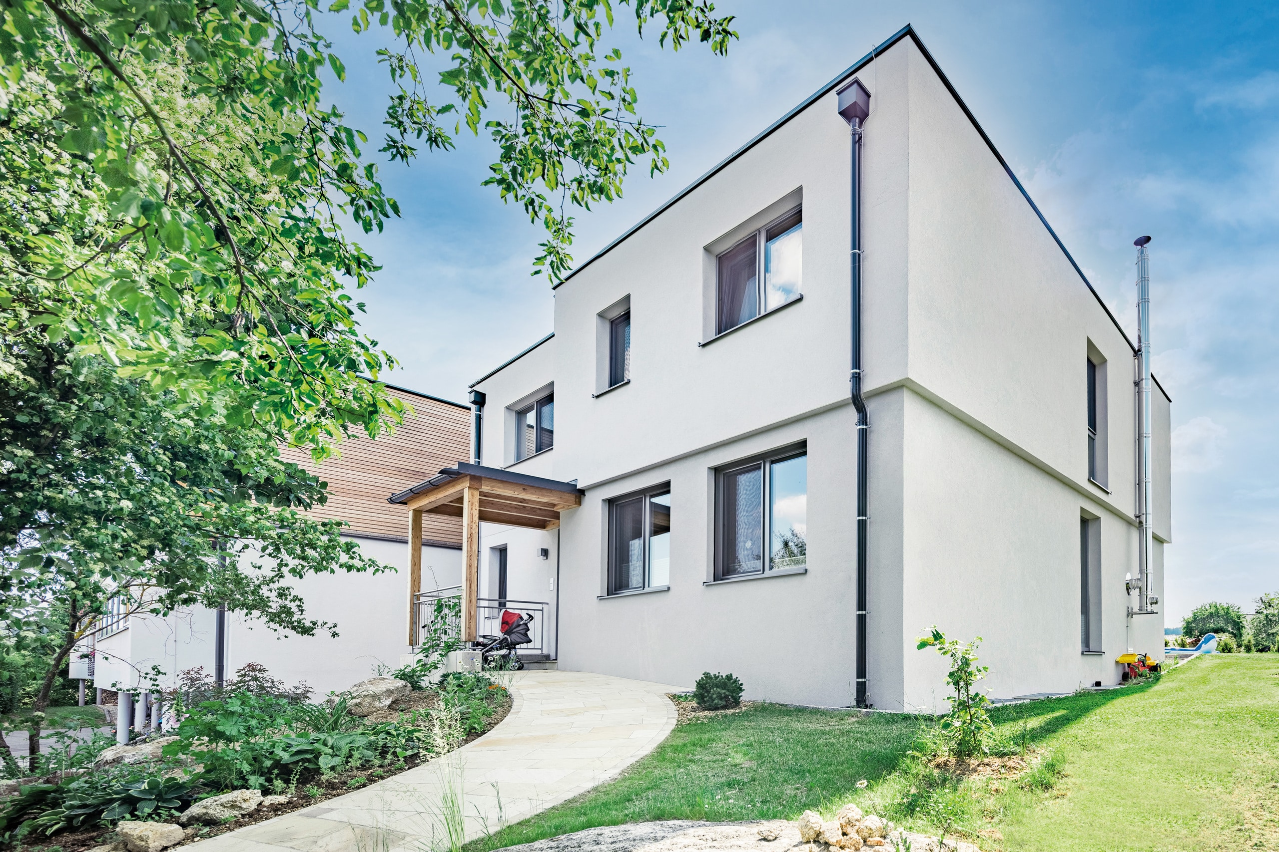 SynthesaGruppe zeigt ein mit Hanf gedämmtes Einfamilienhaus mit gepflastertem Weg zur Haustür, kleinem Vorgarten und Überdachung vom Eingangsbereich.