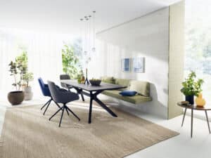 Eine offener Wohnraum mit einem Essbereich mit einer schwebenden Sitzbank in grünem Webstoff auf einer Rückwand aus gebeizter Asteiche und einem Esstisch und Polstersesseln in der Holzausführung tabak, geölt von Tischlerei BAUER.