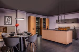 Eine Wohnküche mit Essbereich bestehend aus einem runden Tisch mit 4 Polsterstühlen und einer Inselküche mit Vitrinenschränken in einem Materialmix aus Anthrazit, hellbraun, Holz und Rauchglas von Tischlerei BAUER.
