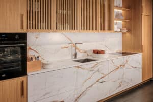 Eine eingekofferte Küchenzeile mit grifflosen Mittelfronten, Arbeitsplatte und Rückwand in weiss-goldener Marmoroptik umrahmt von seitlichen Kästen und Küchenoberschränken mit Holzfronten und Holzlamellen von Tischlerei BAUER.