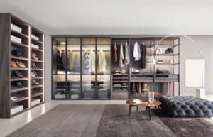 Ein elegantes, modernes Ankleidezimmer mit freiliegendem Teppich, gemütlichem Pouf, Beistelltisch, freistehender Lampe und einem großem Kleiderschranksystem aus Nussbaumholz mit offenen Regalen, Schubladen, indirekter Beleuchtung und Glastüren von BAUER Tischlerei zeigt Sangiacomo.