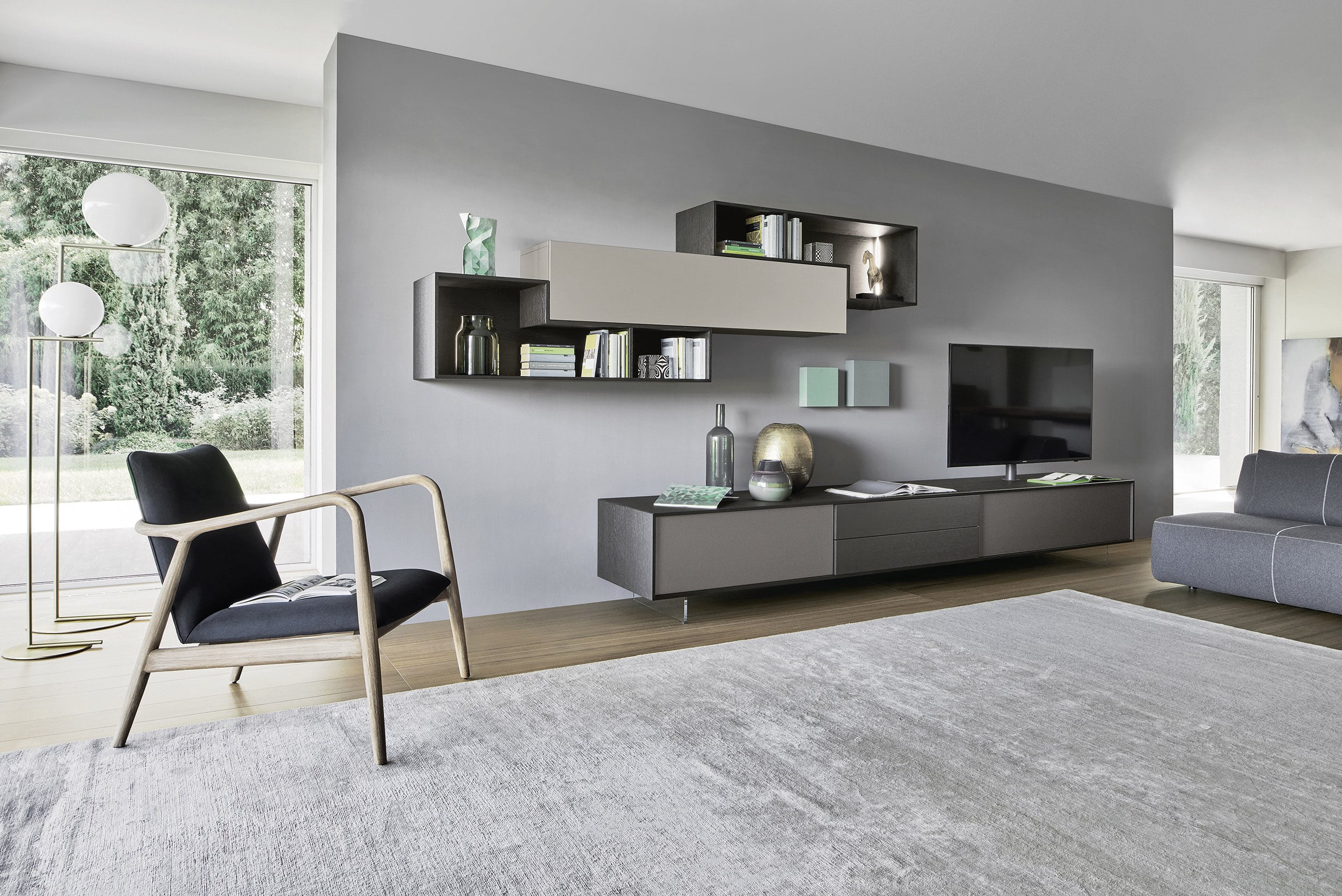Ein zeitgemäßes, stilvolles Wohnzimmer in Grautönen mit freiliegendem Teppich, einem modernen Medien-Board und verschachteltem Wandregal mit Materialmix, Lounge-Chair und kunstvoller Stehleuchte von BAUER Tischlerei zeigt Sangiacomo.