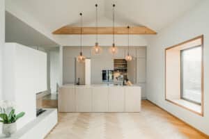 Eine moderne offene Wohnküche mit Kochinsel und eingekofferter Gerätezeile mit versteckter Türe und hellen Holzfronten unter einer Dachschräge mit Holzbalken, Pendelleuchten, Kamin und Sitzfenster von BAUER Tischlerei.
