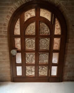 Verspielte Hauseingangstüre mit Rundbogen, gemusterten Glaseinsätzen, passenden Holzeinsätzen und rundem Türknauf mit gedrechseltem Blumenmuster und Sprossen und Rahmen in dunklem Holz von Tischlerei Penninger.