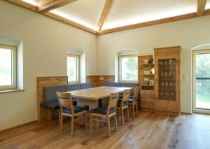 Tischlerei Scharnreithner zeigt einen Essbereich mit Holztisch, Eck-Sitzbank mit stoffbezogener Rückenlehne und Sitzpolster aus Leder, einer Glasvitrine, Kommode, Regale und Wandverkleidung aus Holz.