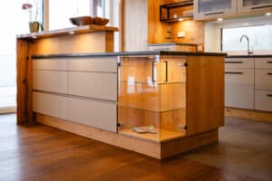 Eine moderne Küche mit Kochinsel und integrierter beleuchteter Glasvitrine in klarer Linienführung und elegantem Holz‑Glas‑Kontrast von Tischlerei Scharnreitner.