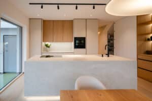 Eine moderne, offene Wohnküche mit beigen grifflosen Fronten, Holzakzenten in Form eines Oberschranks, Schrankwand mit Schiebetüren und Holzinnenleben und einer Kochinsel in passender Beton bzw. Steinoptik in minimalistischem Design von Tischlerei Scharnreitner.