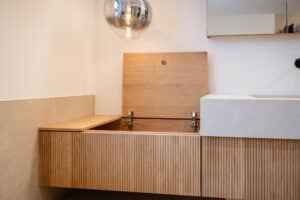 Ein modernes Badezimmer mit schwebendem Doppelwaschtisch in beiger Steinoptik, Pendelleuchte und Waschtischunterbau aus Holz mit moderner geriffelter Oberfläche und einer Truhenöffnung als zusätzliche Aufbewahrungsmöglichkeit von Tischlerei Scharnreitner.