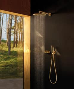VOLA präsentiert eine große runde Regendusche, eine stabförmige Handdusche und zwei Reglern aus mattem Gold an der Wand montiert.