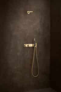 VOLA präsentiert eine stabförmige Kopfdusche mit beidseitigen Düsen, Umschaltgriffe und Handdusche mit dunkelblauem Griff aus mattem Gold.