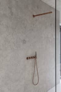 VOLA zeigt eine Dusche mit grau marmorierten Fliesen und kupferfarbenen Dusch-Armatur mit stabförmiger Kopf- und Handdusche und seperaten Drehschaltern.