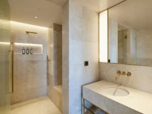 VOLA zeigt ein Badezimmer eines Hotelzimmers mit Marmor-Waschbecken mit goldenen Armaturen und eine Dusche mit stabförmiger Kopf- und Handdusche in gold.